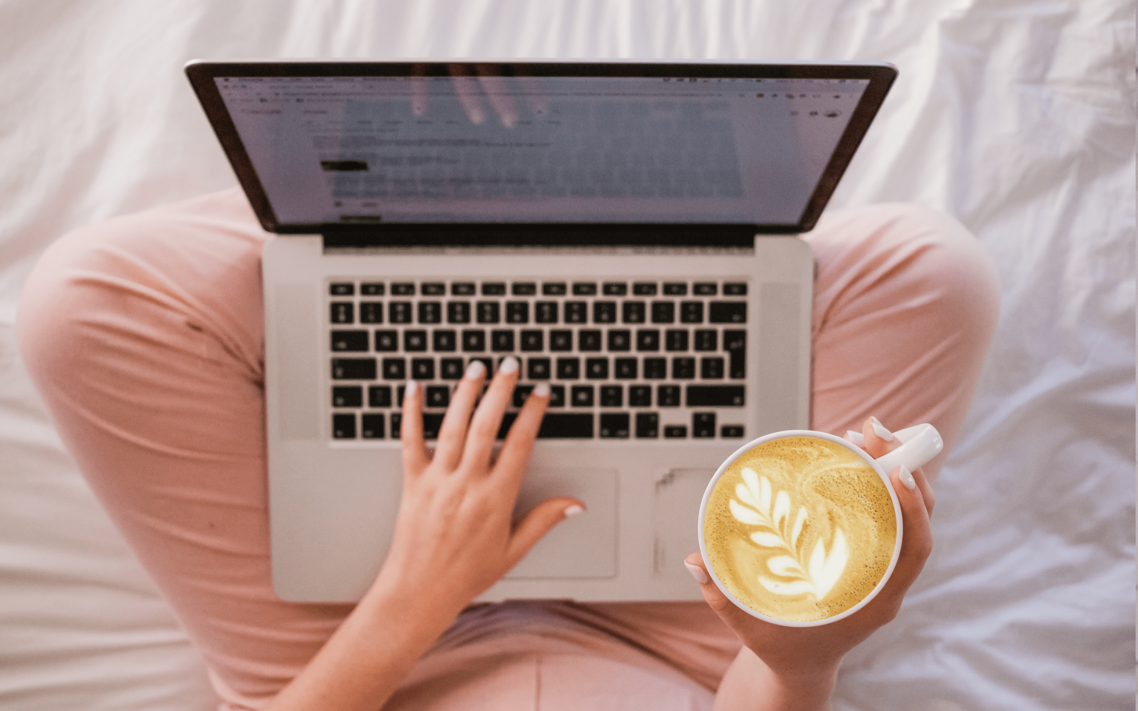 A woman with white painted fingernails sits on a duvet crossed legged. Her laptop is resting on her knees, while one hand types on her MacBook and the other holds a cup of Latte style coffee.